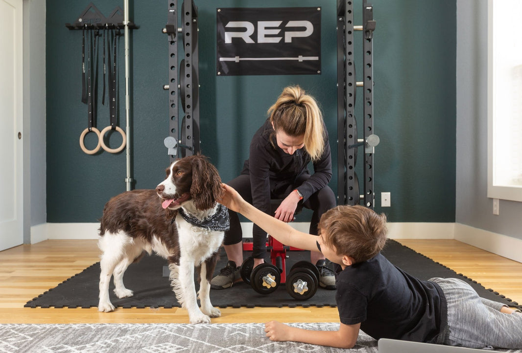 A family hangs out in their home gym: a boy pets his dog while the mom gets ready to lift.