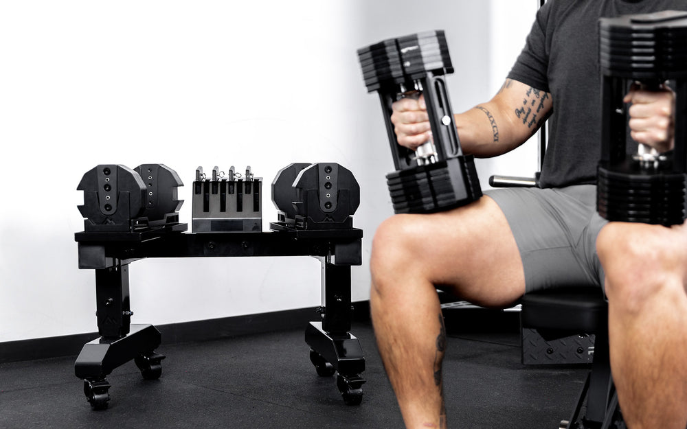 Athlete working out with REP® x PÉPIN™ adjustable dumbbells in front of a metallic black REP® Adjustable Dumbbell Stand