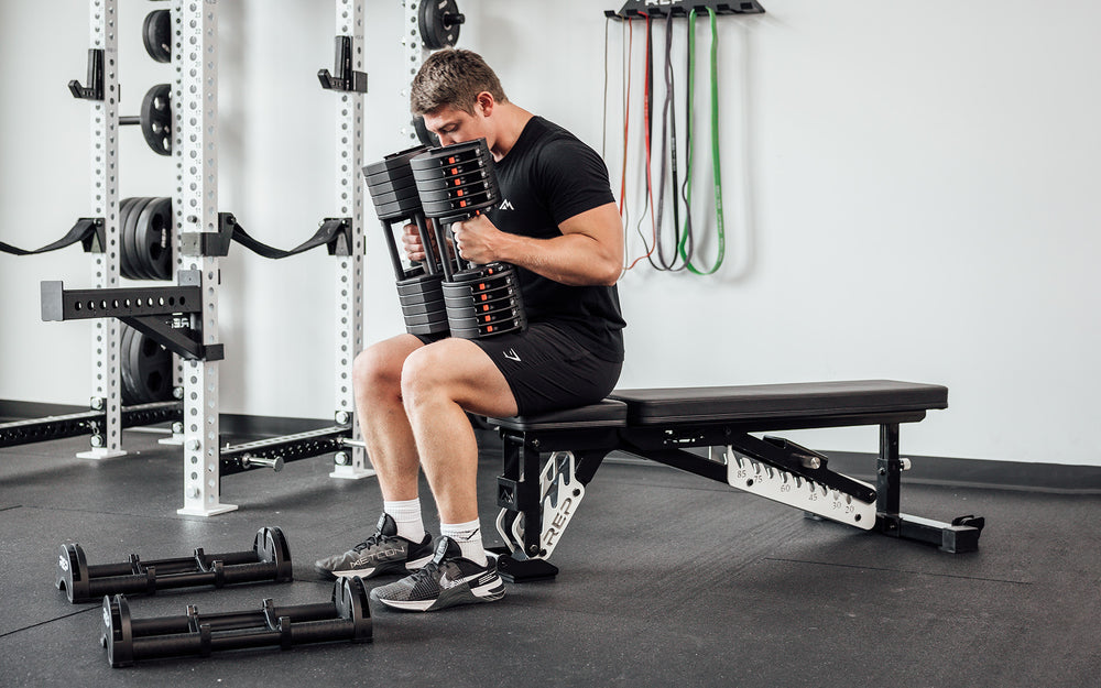 Male lifter holding a pair of 60lb REP Fitness QuickDraw Adjustable Dumbbells and preparing to lay back on a bench to perform bench presses.