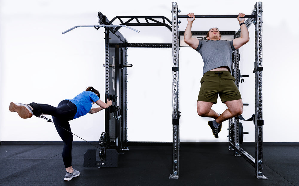 1 person doing pull-ups in a PR-5000 rack while another person is using the low row for leg exercises on the Adonis™ Cable Tower.