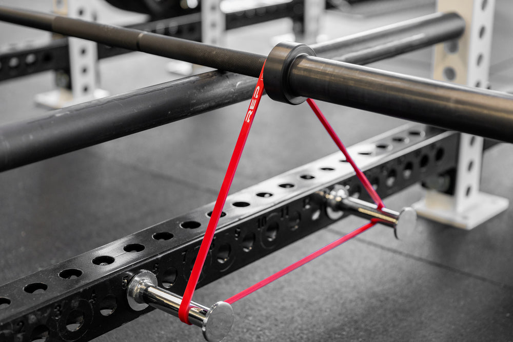 Empty barbell on pin-pipe safeties with red short resistance bands attached using band pegs for banded bent over rows.