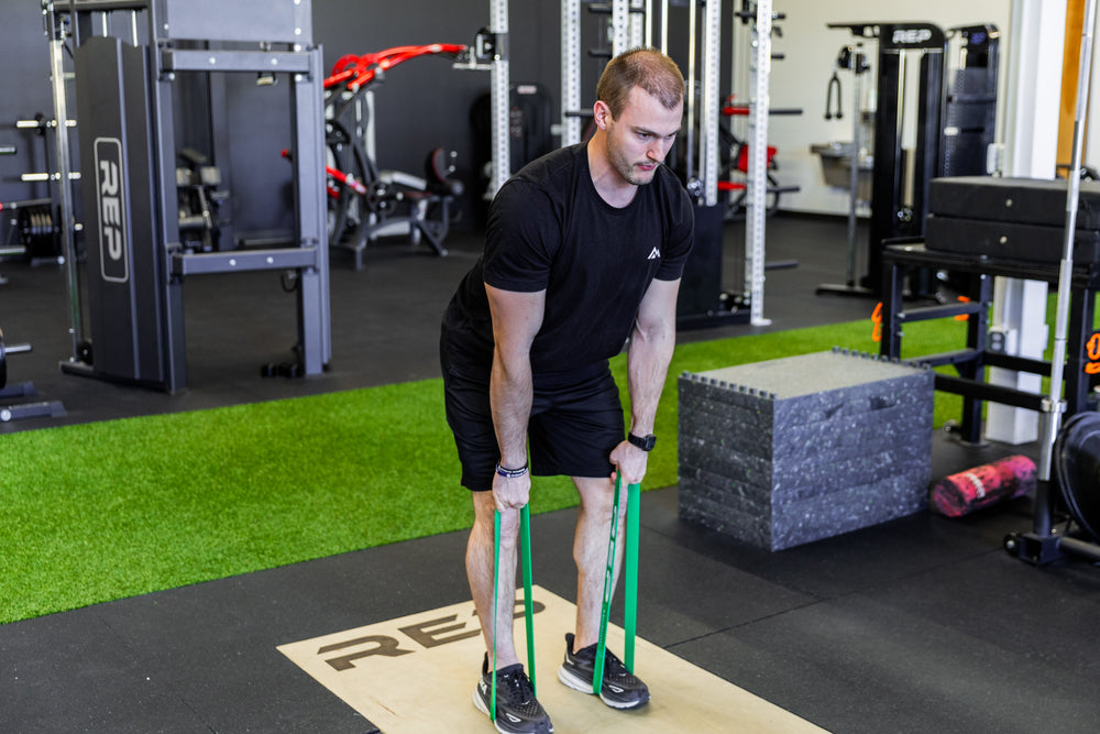 Lifter using green short resistance bands around his feet to perform banded deadlifts.