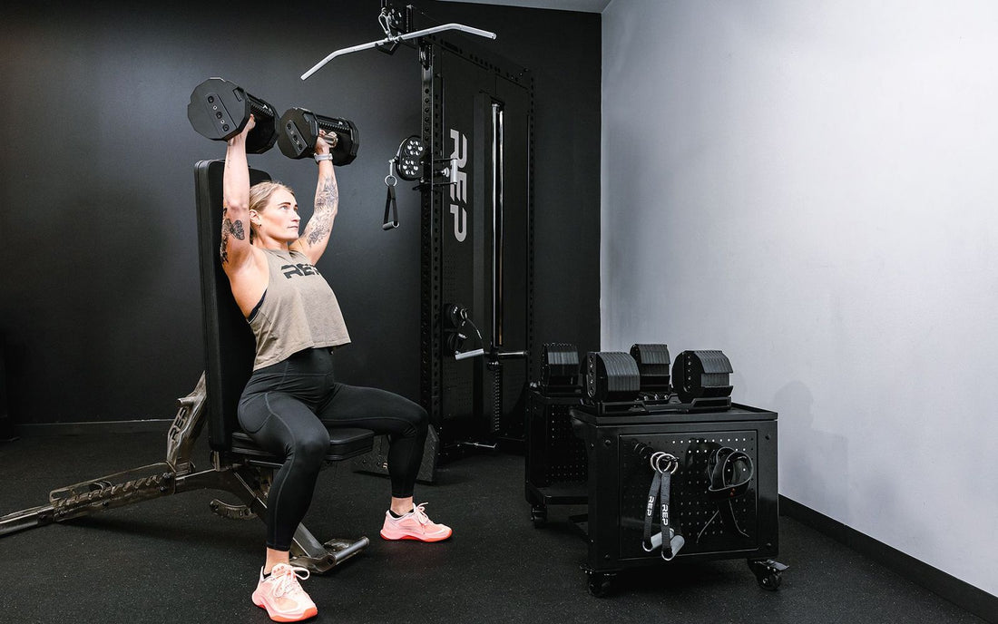 An athlete shoulder pressing the REP x PÉPIN FAST Series Adjustable Dumbbells next to the REP Dumbbell Storage Cart.