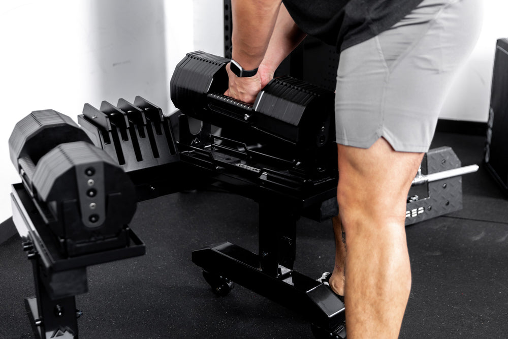 Athlete picking up REP® x PÉPIN™ adjustable dumbbells from a metallic black REP® Adjustable Dumbbell Stand from one side