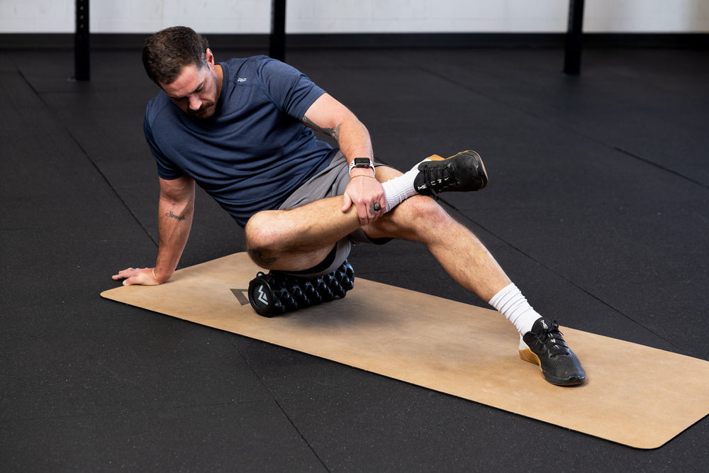 Man using High Density Foam Roller to roll out glutes on a cork yoga mat in a gym setting.