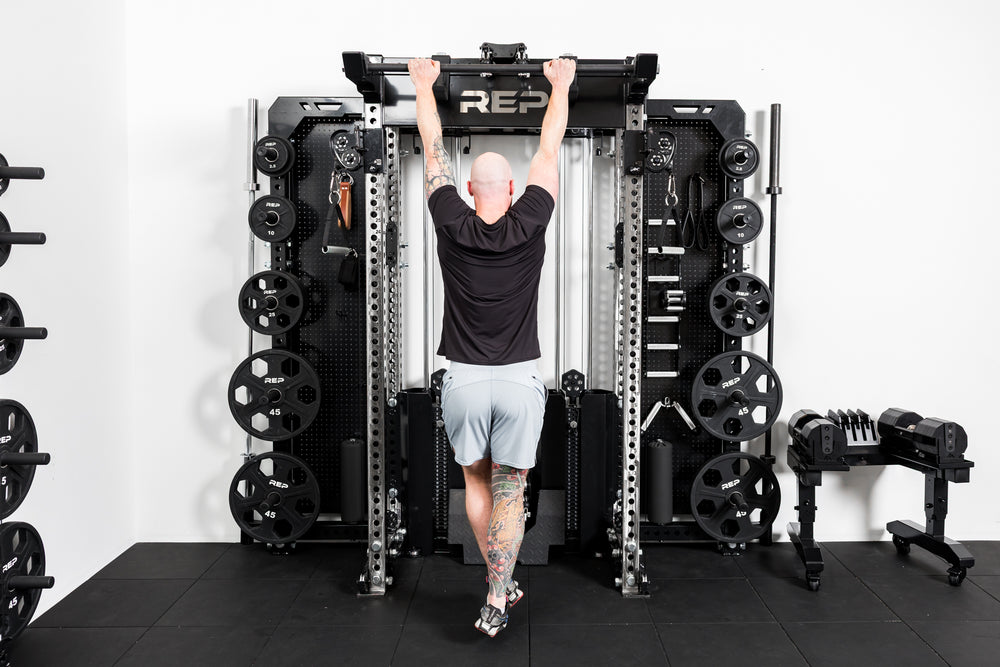 Person using a Rep Fitness power rack in a gym setting with the standard Logo Plate Crossmember 2.0 shown - black with laser-cut "REP".
