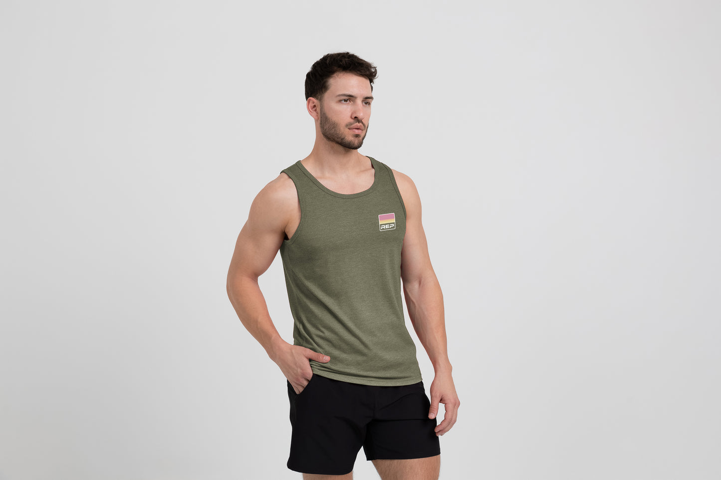 Man wearing an olive Men's Rise Tri-Blend Tank and shorts on a white background