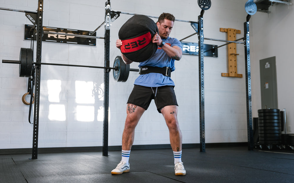 Athlete performing a ground to over the shoulder lift with a REP Stone Sandbag while wearing a REP Nylon Lifting Belt.