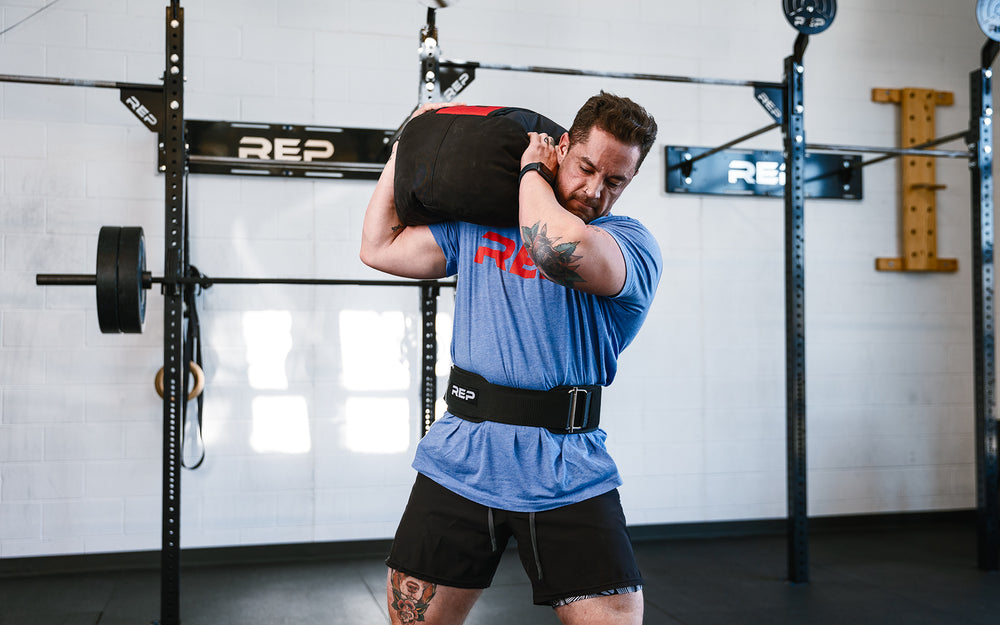 Athlete performing a ground to over the shoulder lift with a REP Stone Sandbag while wearing a REP Nylon Lifting Belt.