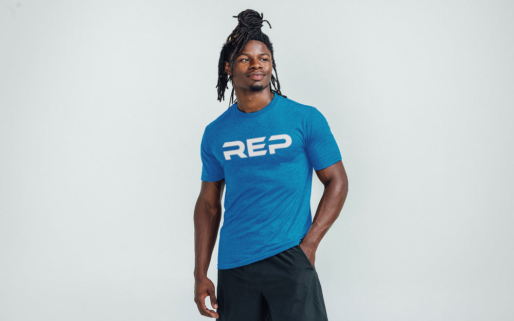 Model wearing the REP Fitness Men's Daily Driver 2.0 Tri-Blend Crew T-Shirt in Heather Royal Blue with a White graphic
