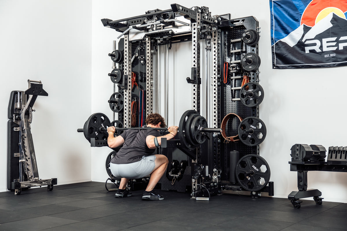 Person performing back squat off the front of a Wall-Mounted Ares™ 2.0 Rack in a gym setting.