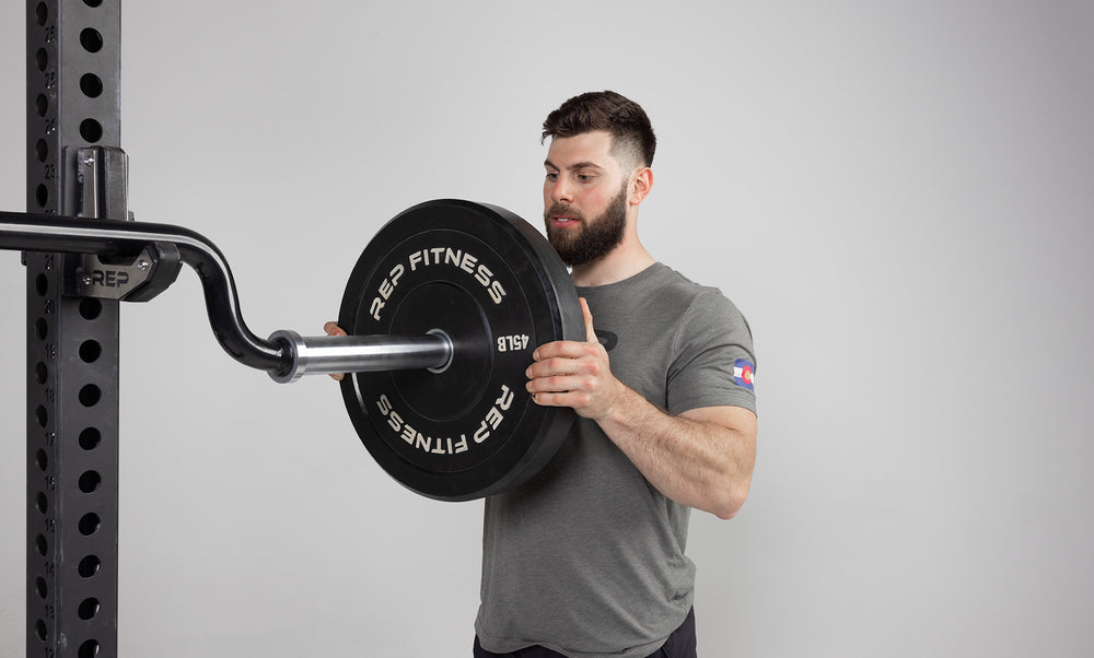 Lifter sliding a bumper plate on the hard chrome sleeve of a racked Safety Squat Bar.