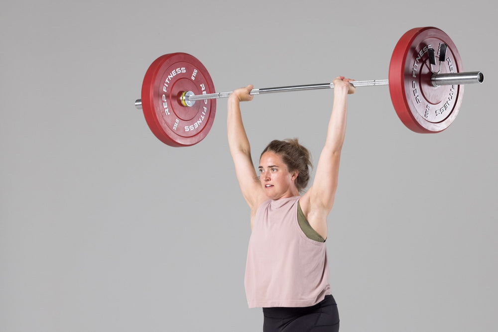 Lifter in finish position of a split jerk with a barbell overhead loaded with a pair of red 55lb colored bumper plates.