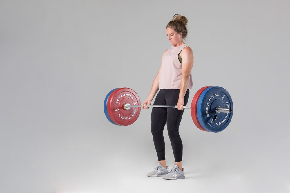 Lifter at the top position of a deadlift using a barbell loaded with a pair of red 55lb and blue 45lb colored bumper plates.