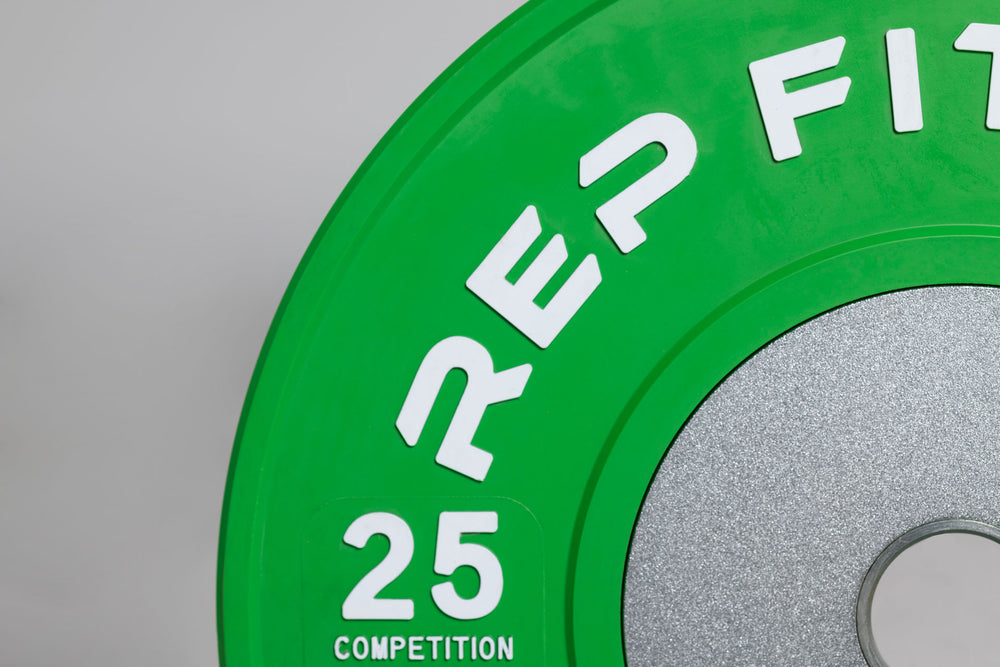 Close-up view of a green 25lb competition bumper plate showing zinc-coated steel disc insert and raised white "REP FITNESS" and weight marking lettering.