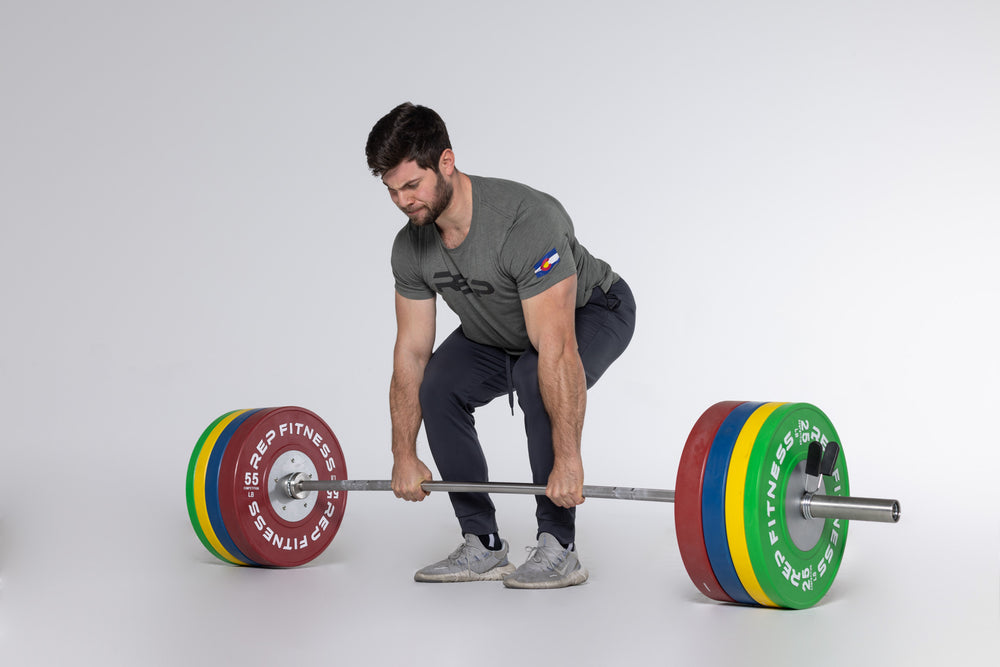 Lifter in the set-up position getting ready to perform a deadlift with a barbell loaded with a pair of red 55, blue 45, yellow 35, and green 25lb competition bumper plates.