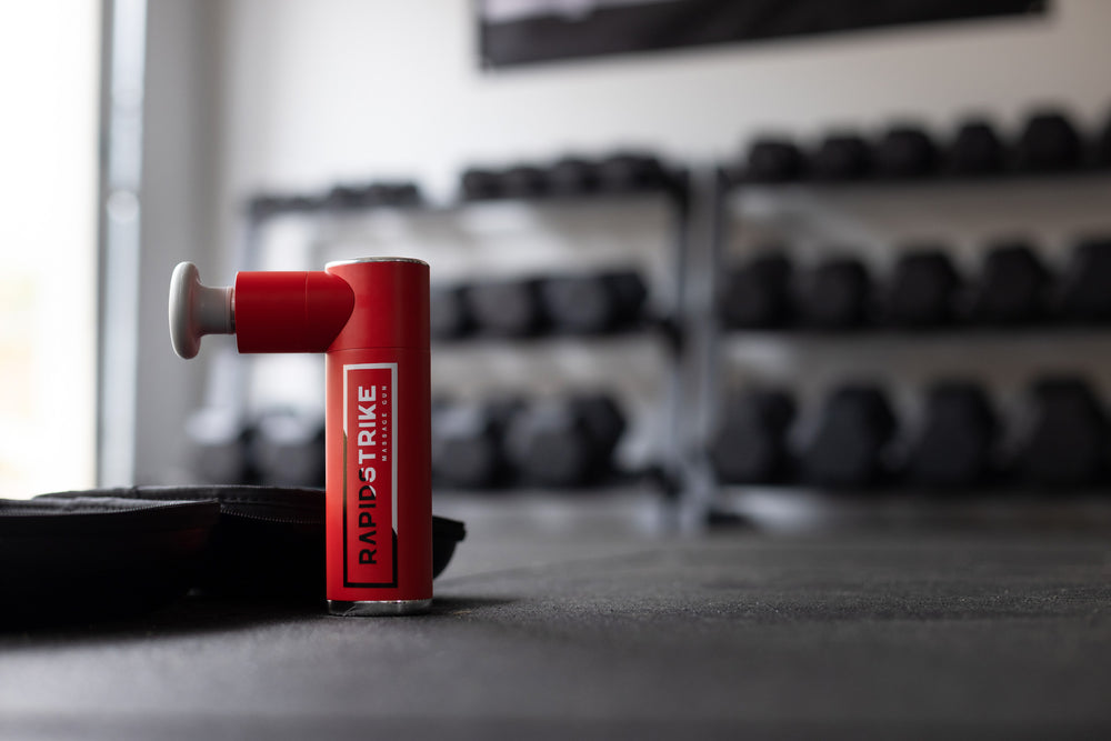 Close-up view of the Rapidstrike massage Gun standing up by its carrying case on the floor of a home gym.