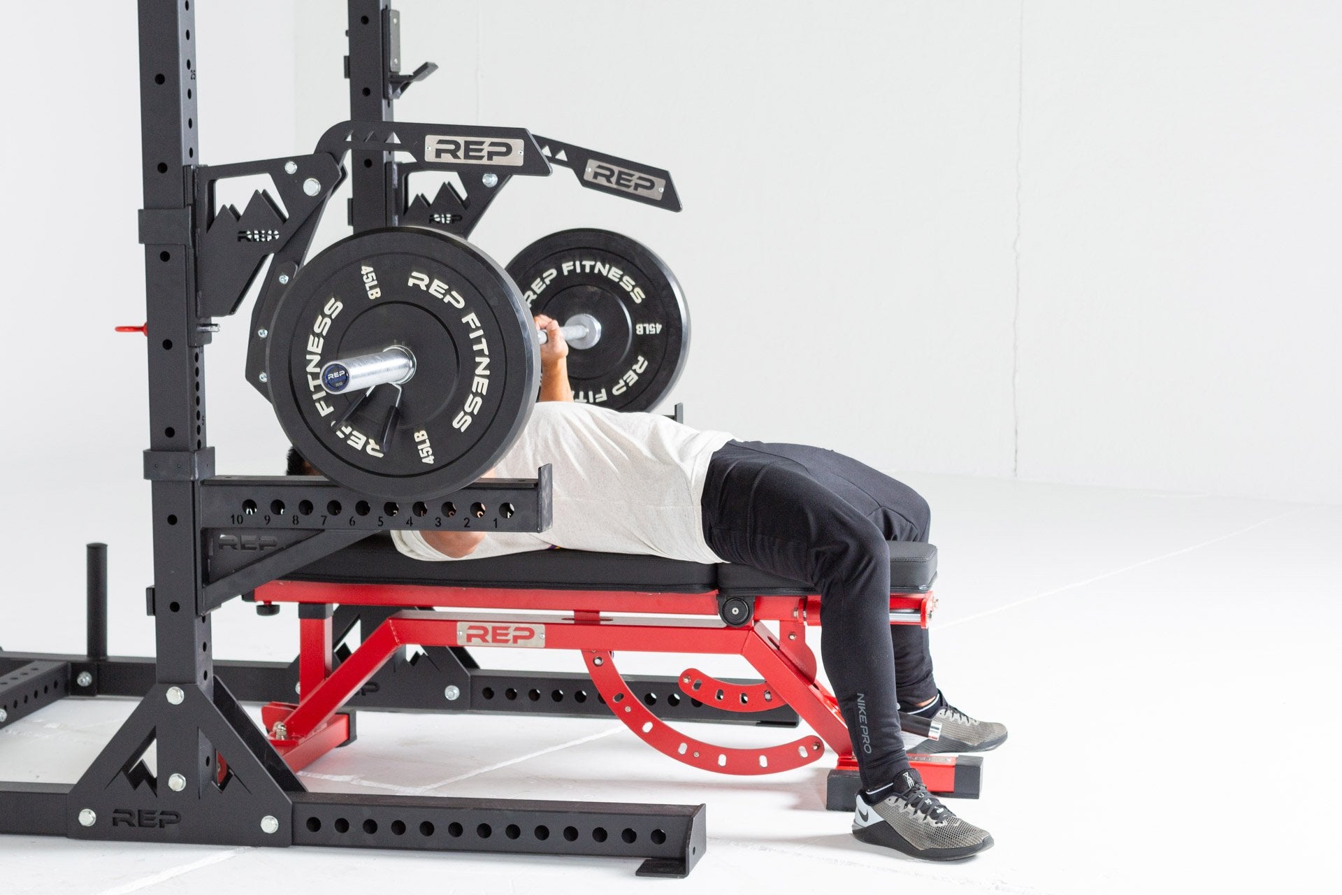 Lifter lying on an AB-5000 Bench inside a REP Squat Rack with REP Monolift Arms and Spotter Arms performing a bench press with a barbell loaded with a pair of 45lb Black Bumper Plates.