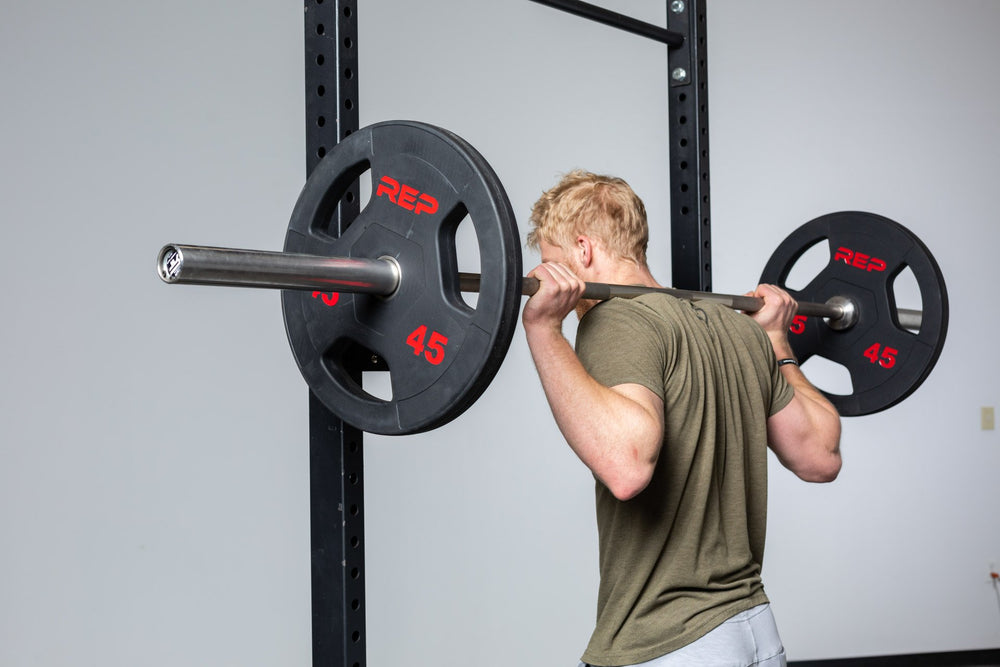 Lifter standing with a barbell loaded with a pair of 45lb Rubber Coated Olympic Plates in the back rack position outside of a squat rack.